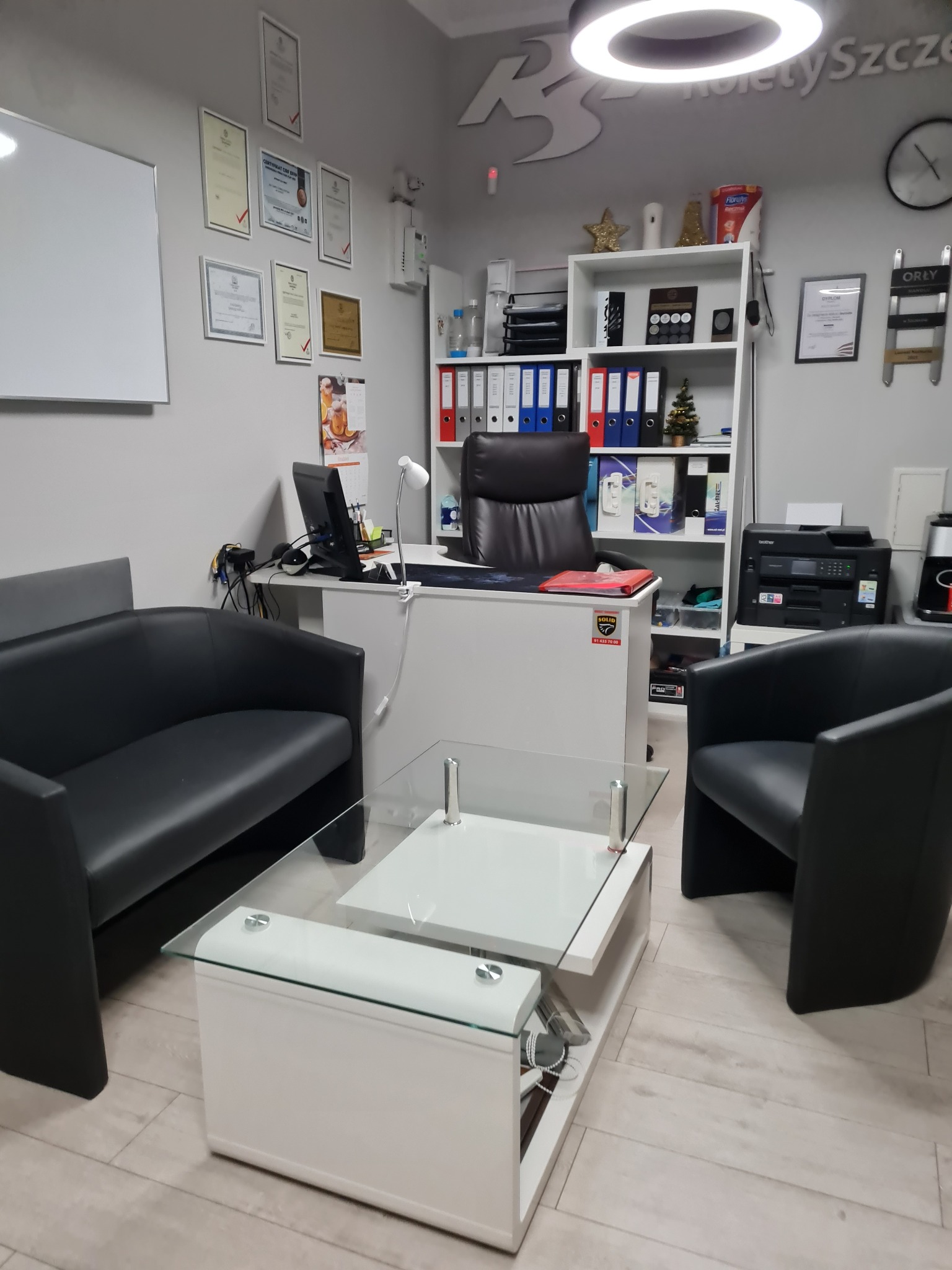 Wnętrze biura firmy z roletami: białe biurko, czarne fotele, szklany stolik, regał z dokumentami i nagrodami, dyplomy na ścianie, logo firmy na ścianie.