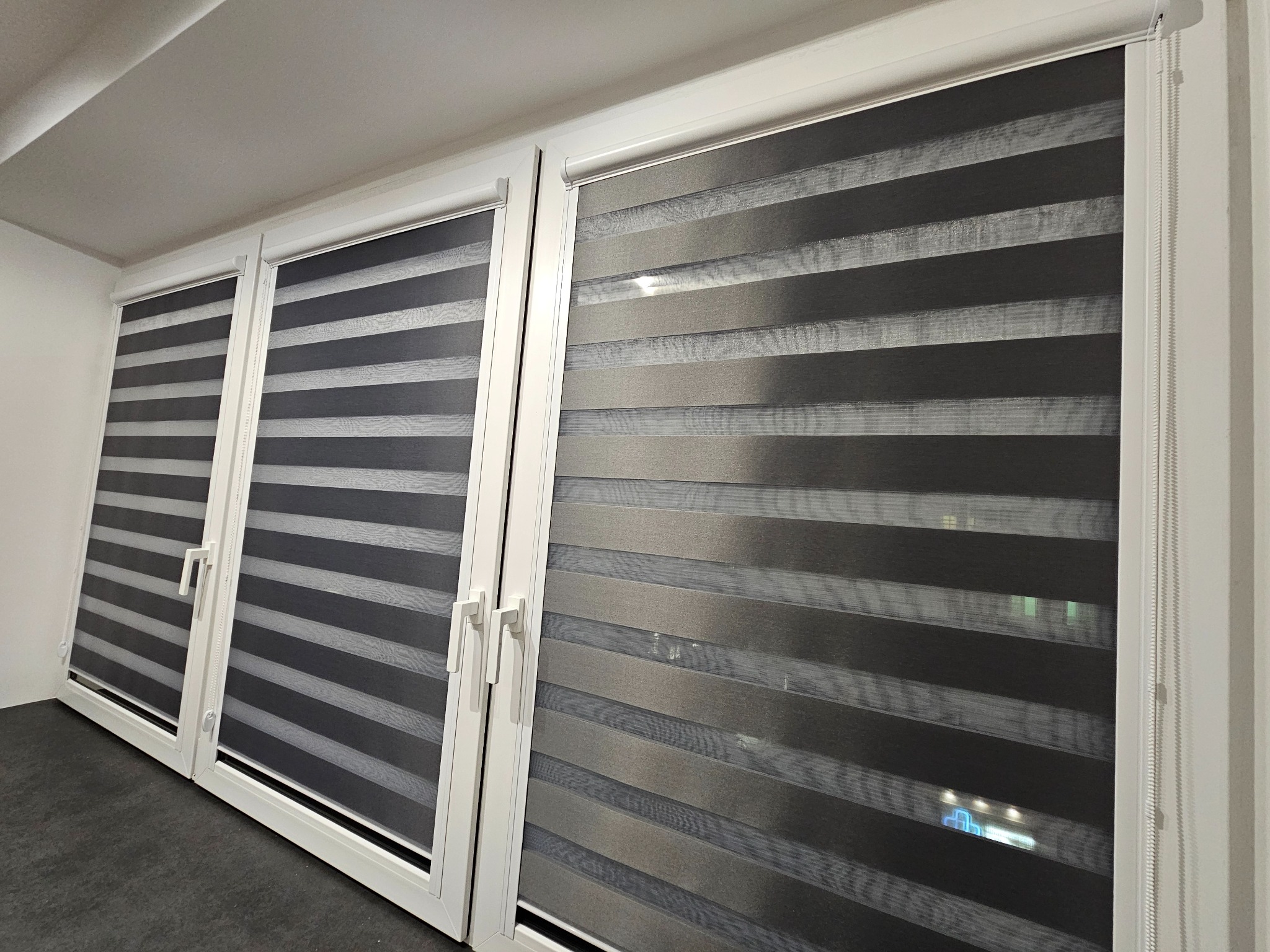 Trzy okna z zamontowanymi roletami dzień noc w odcieniach szarości, widoczne uchwyty okienne i mechanizm rolety po prawej stronie. Nowoczesny design.