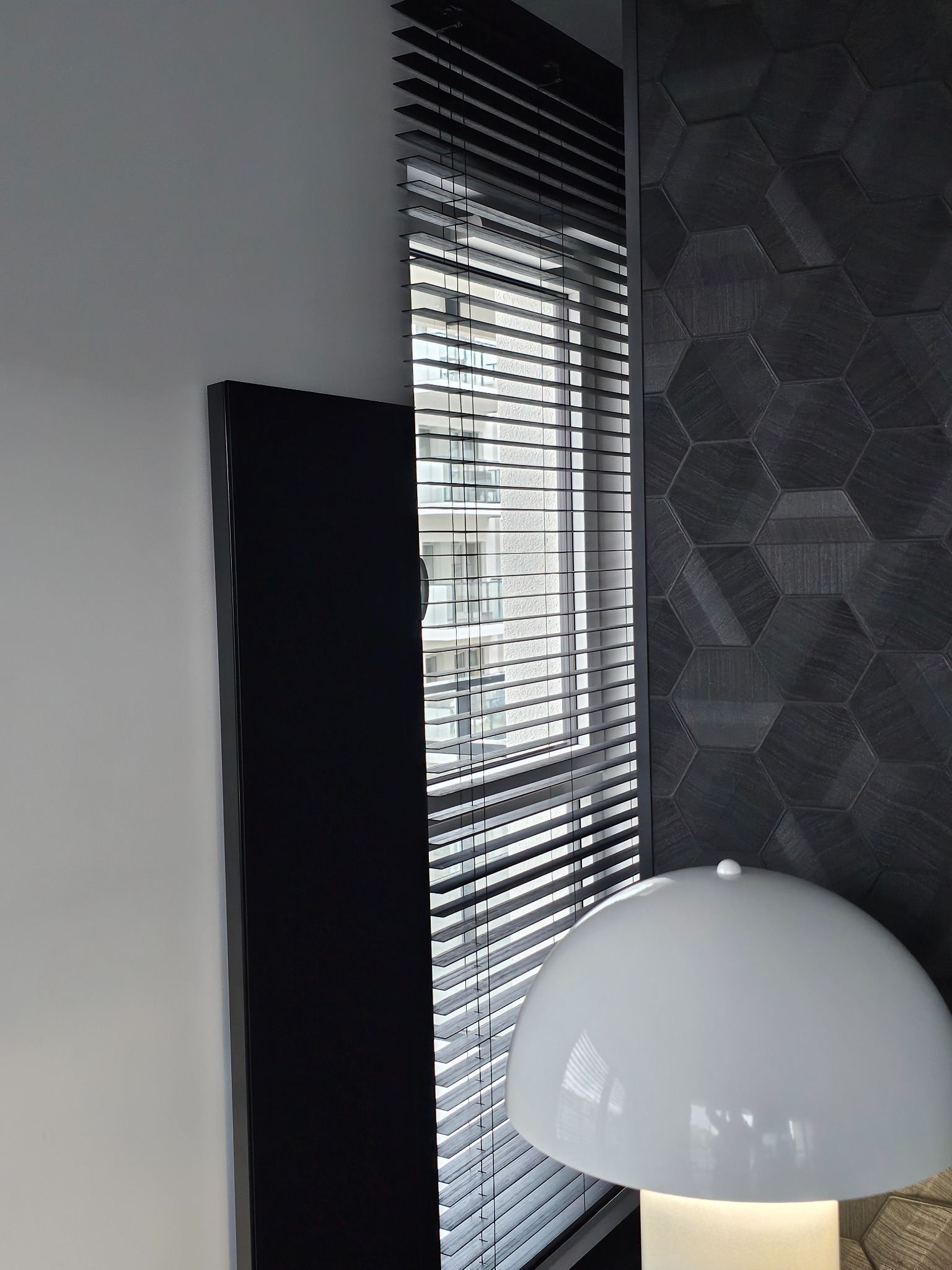Ciemne żaluzje poziome w oknie, z widokiem na budynek. Obok ściana z heksagonalnymi płytkami i lampa. Minimalistyczny design wnętrza.