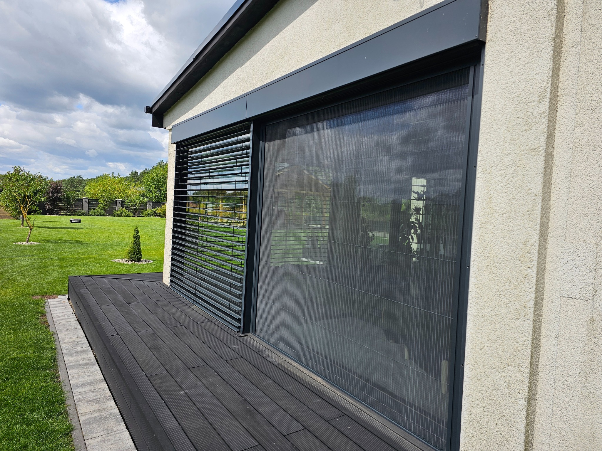 Nowoczesna moskitiera przesuwna w ciemnej ramie, zamontowana na oknie tarasowym. Obok widoczne żaluzje poziome w kolorze antracytowym i drewniany taras.