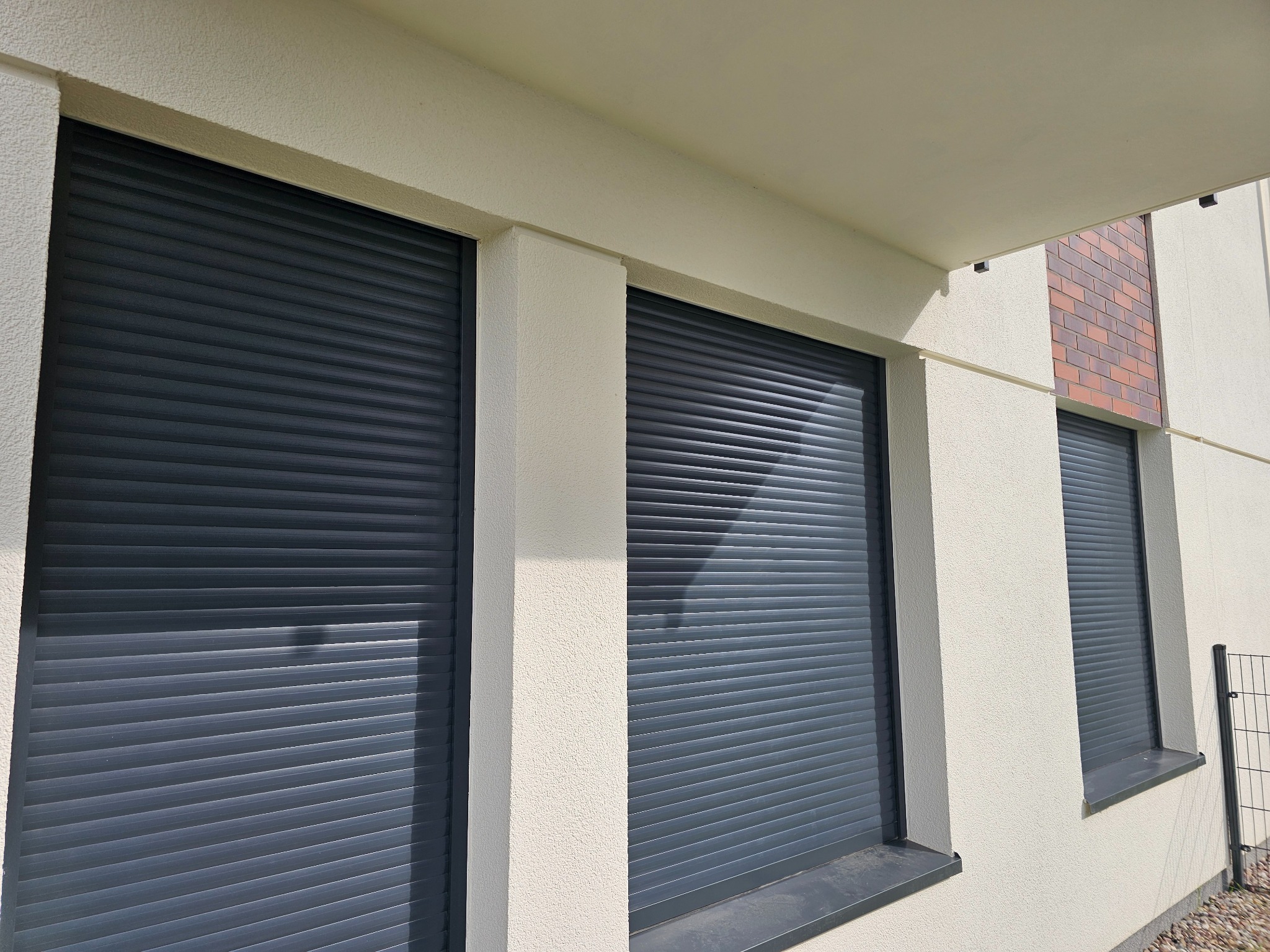 Elewacja budynku z trzema oknami zabezpieczonymi szarymi roletami zewnętrznymi, widoczne fragmenty ceglanej dekoracji i ogrodzenia.