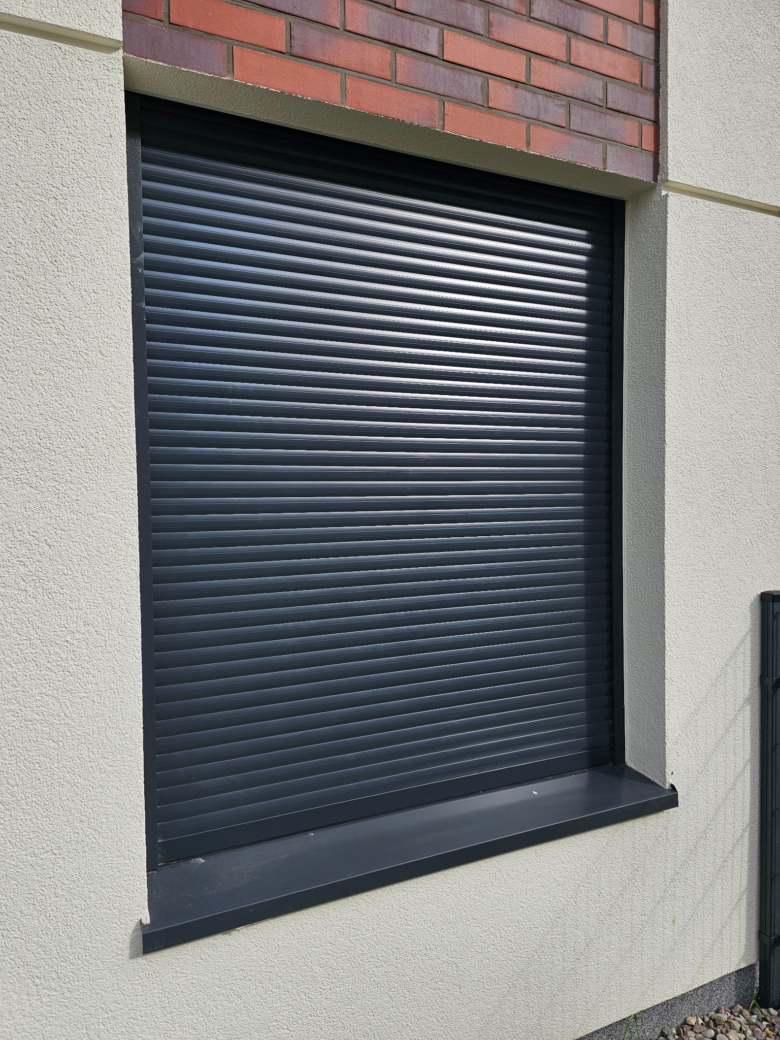 Zamknięta, grafitowa roleta zewnętrzna na oknie budynku z elewacją w jasnym odcieniu i fragmentem muru z cegły. Widoczne poziome lamele rolety i parapet.