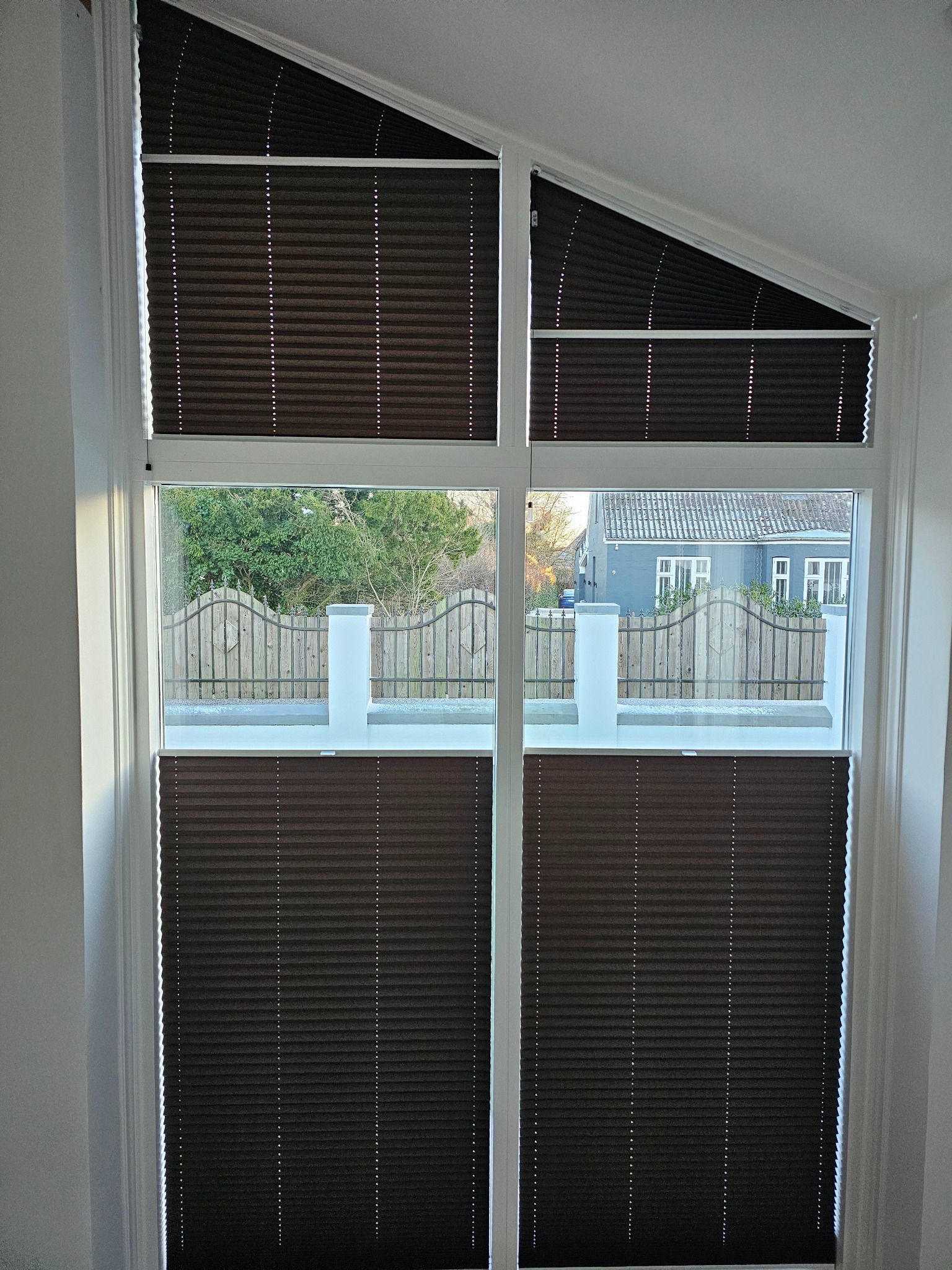 Ciemnobrązowe plisy okienne zamontowane w oknie o nieregularnym kształcie. Widok na zewnątrz, gdzie widać ogrodzenie i dom. Plisy dopasowane do nietypowego kształtu okna.