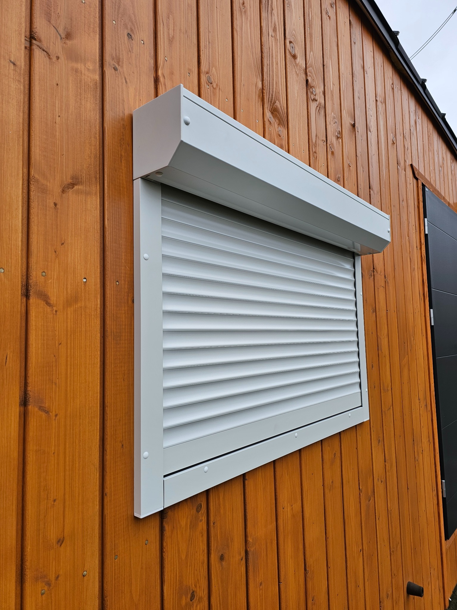 Biała roleta zewnętrzna częściowo opuszczona na oknie drewnianego budynku. Widoczne poziome lamele i kaseta rolety. Ujęcie z boku, podkreślające detale montażu.