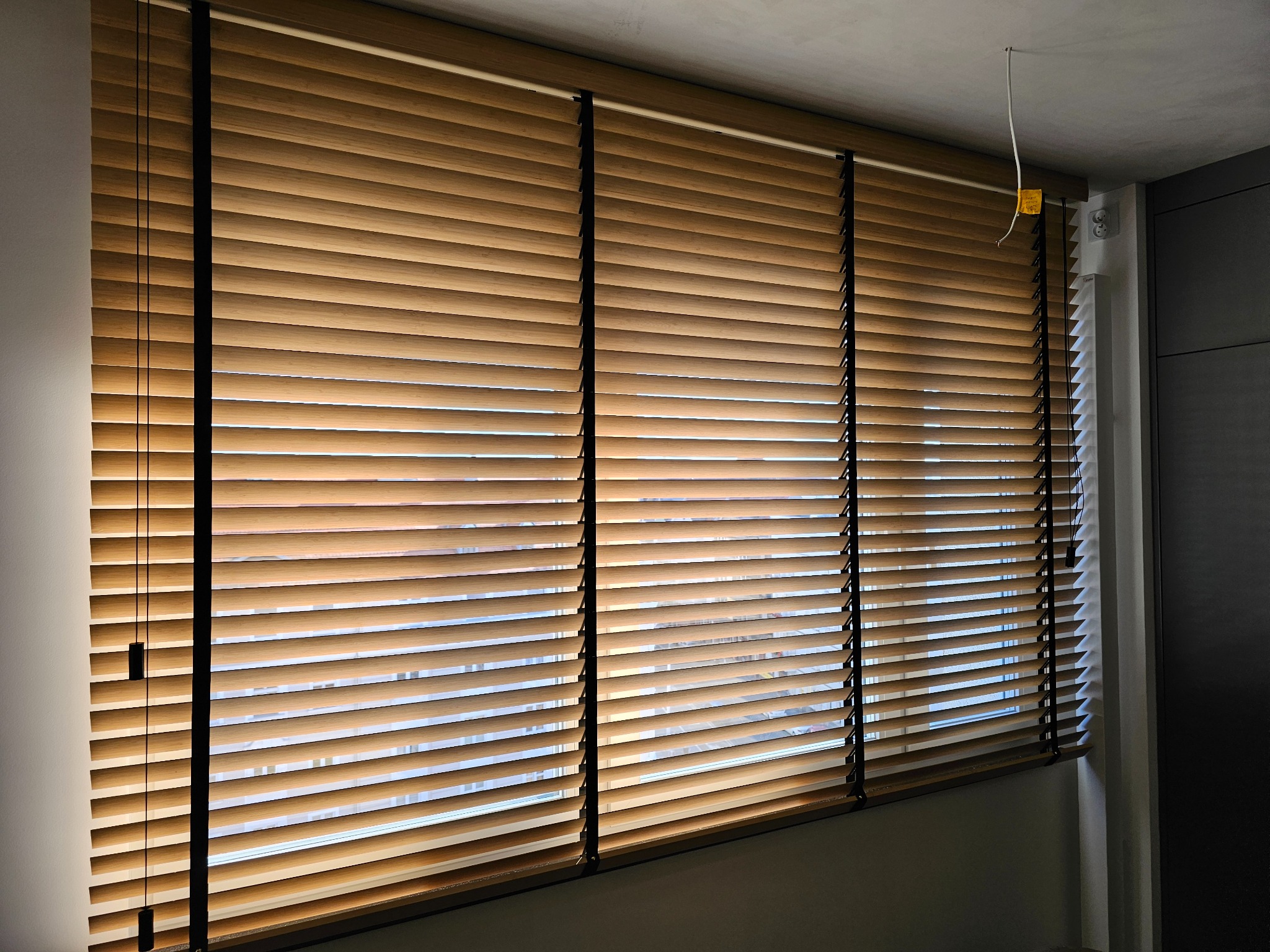 Zamknięte żaluzje drewniane w ciepłym odcieniu brązu, z czarnymi sznurkami, zamontowane w oknie. Widoczne elementy sufitu i ściany w neutralnych kolorach.