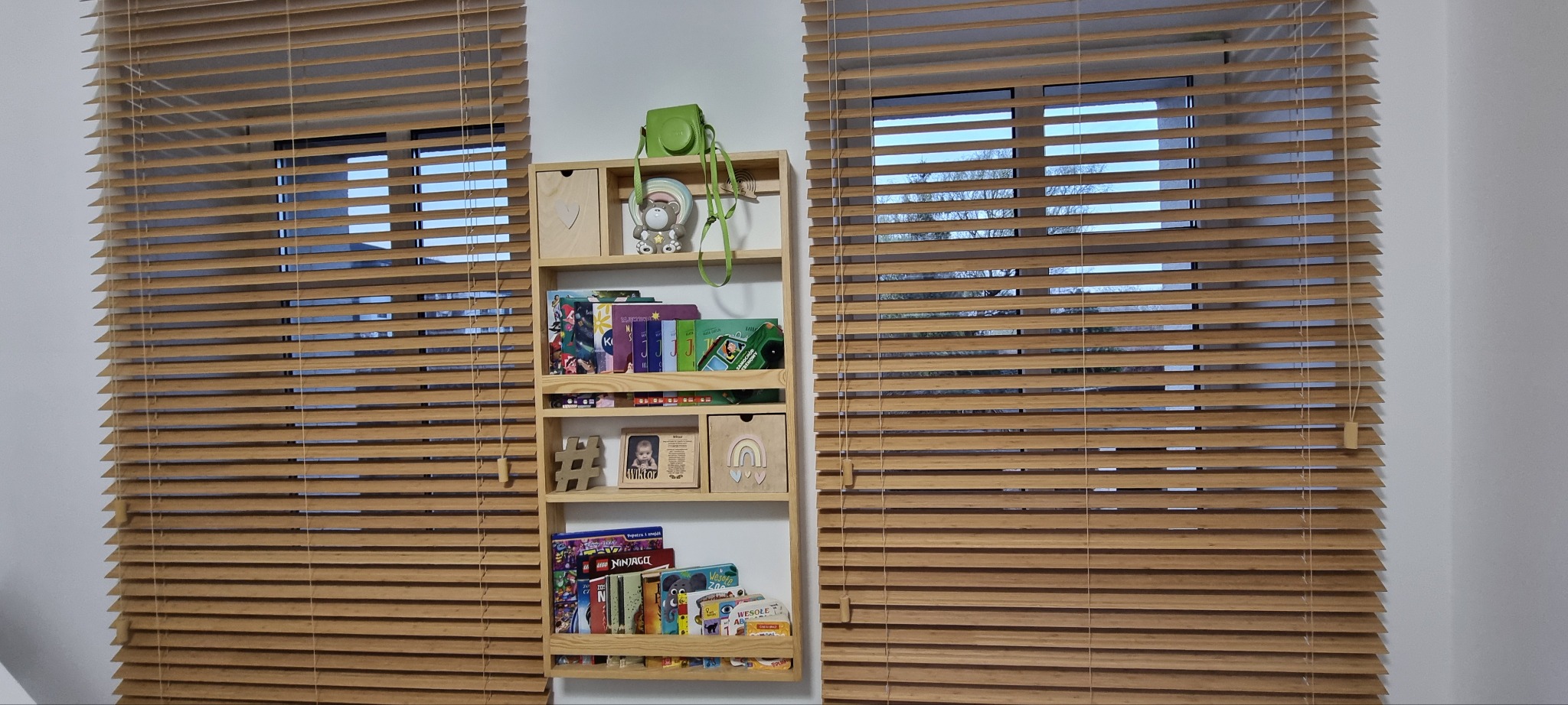 Drewniane żaluzje poziome w ciepłym odcieniu drewna, zamontowane w oknach pokoju dziecięcego, z półką na książki i zabawki pośrodku. Naturalne światło dzienne.