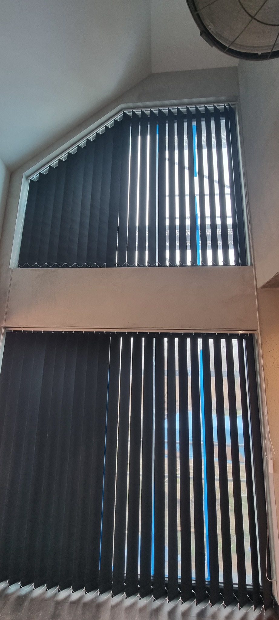 Czarne, pionowe żaluzje w dwóch oknach o nietypowym kształcie. Górne okno trójkątne, dolne prostokątne. Widoczne sznurki do regulacji. Minimalistyczny design.