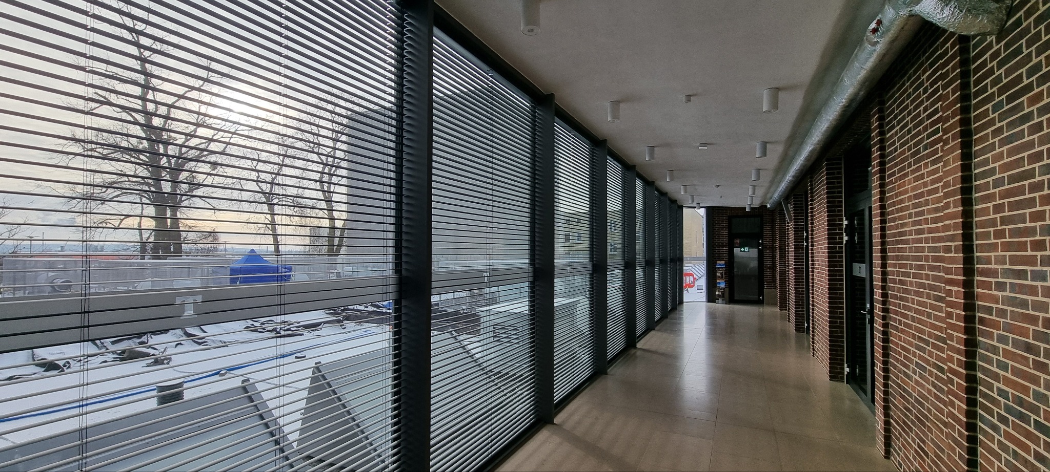 Wnętrze korytarza z rzędem okien przesłoniętych aluminiowymi żaluzjami w kolorze grafitowym. Widok na zewnątrz z drzewami i elementami miejskiego krajobrazu.