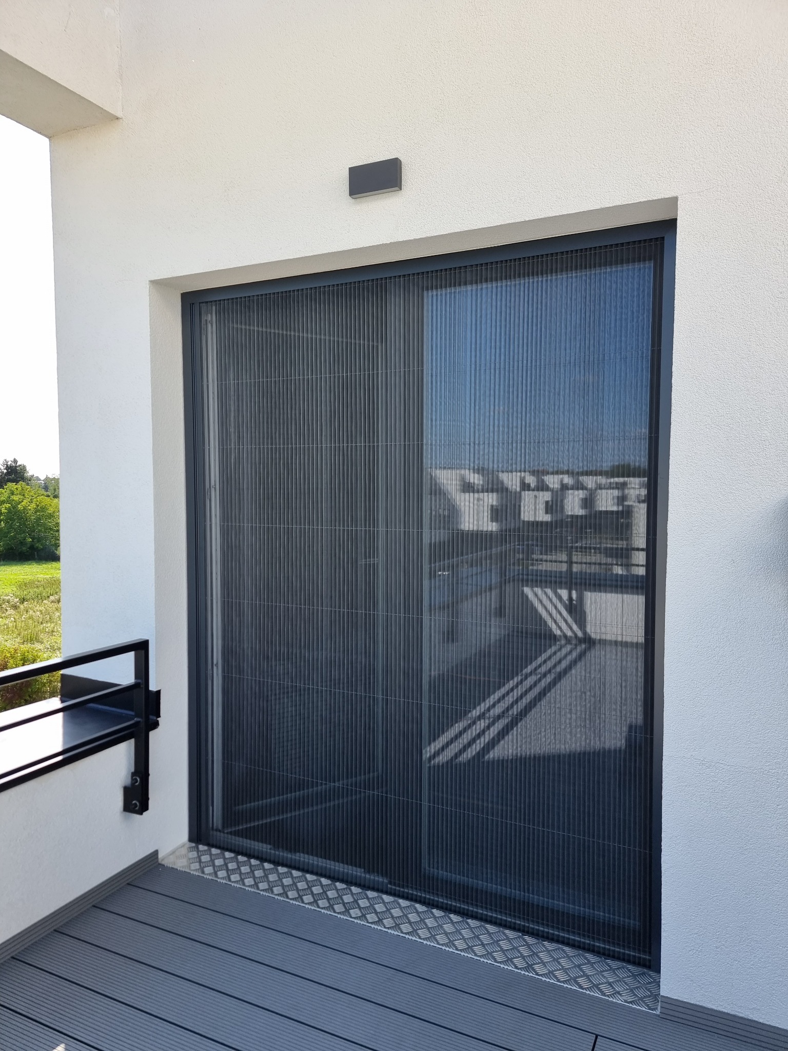 Zamknięta, plisowana moskitiera w grafitowej ramie, zamontowana na balkonie z szarą podłogą. Widok z zewnątrz, w tle zabudowa i zieleń.