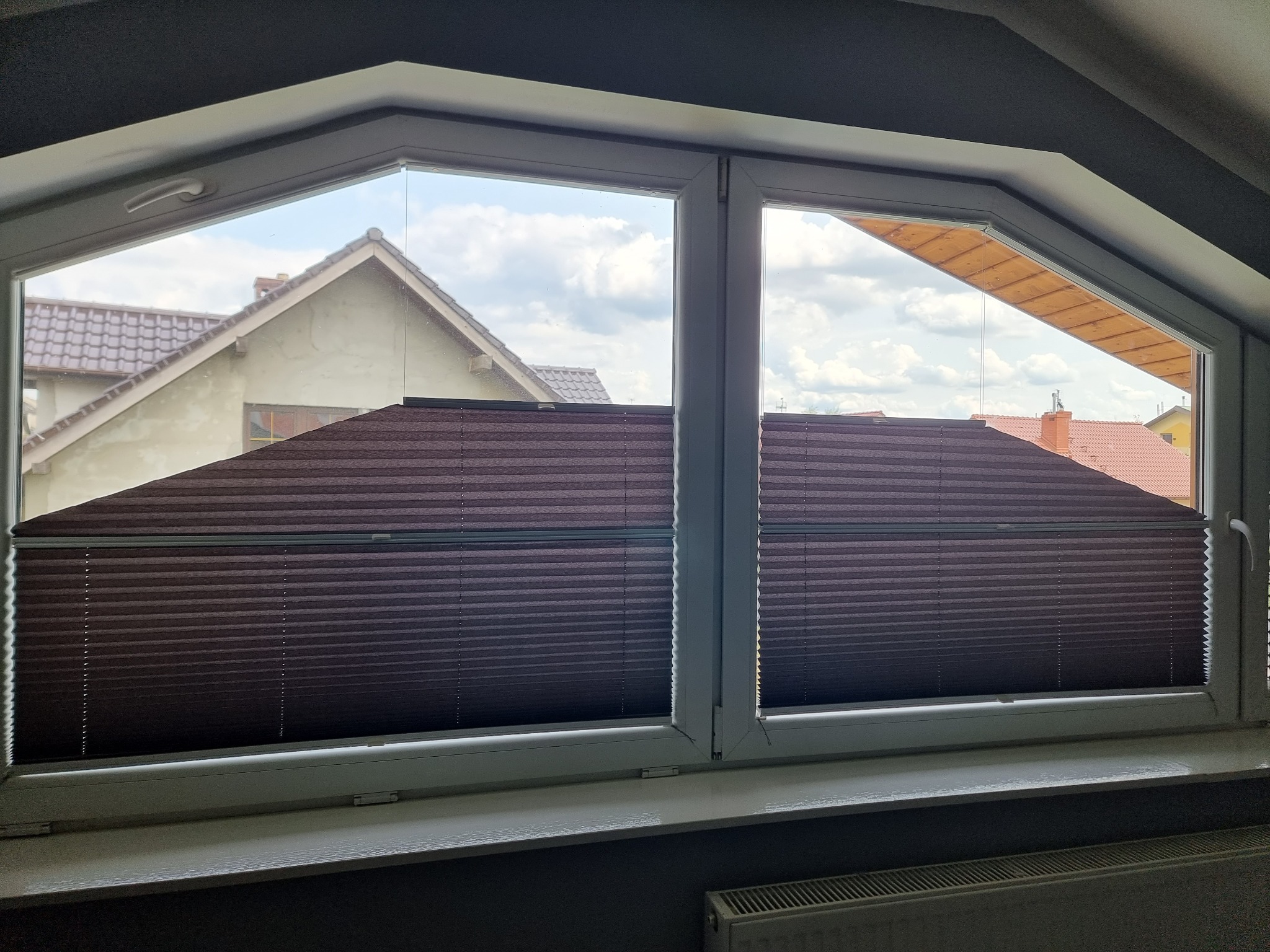 Brązowe plisy w oknie o nietypowym kształcie, widok na dom i niebo. Plisy częściowo zasłonięte, z widoczną strukturą materiału.