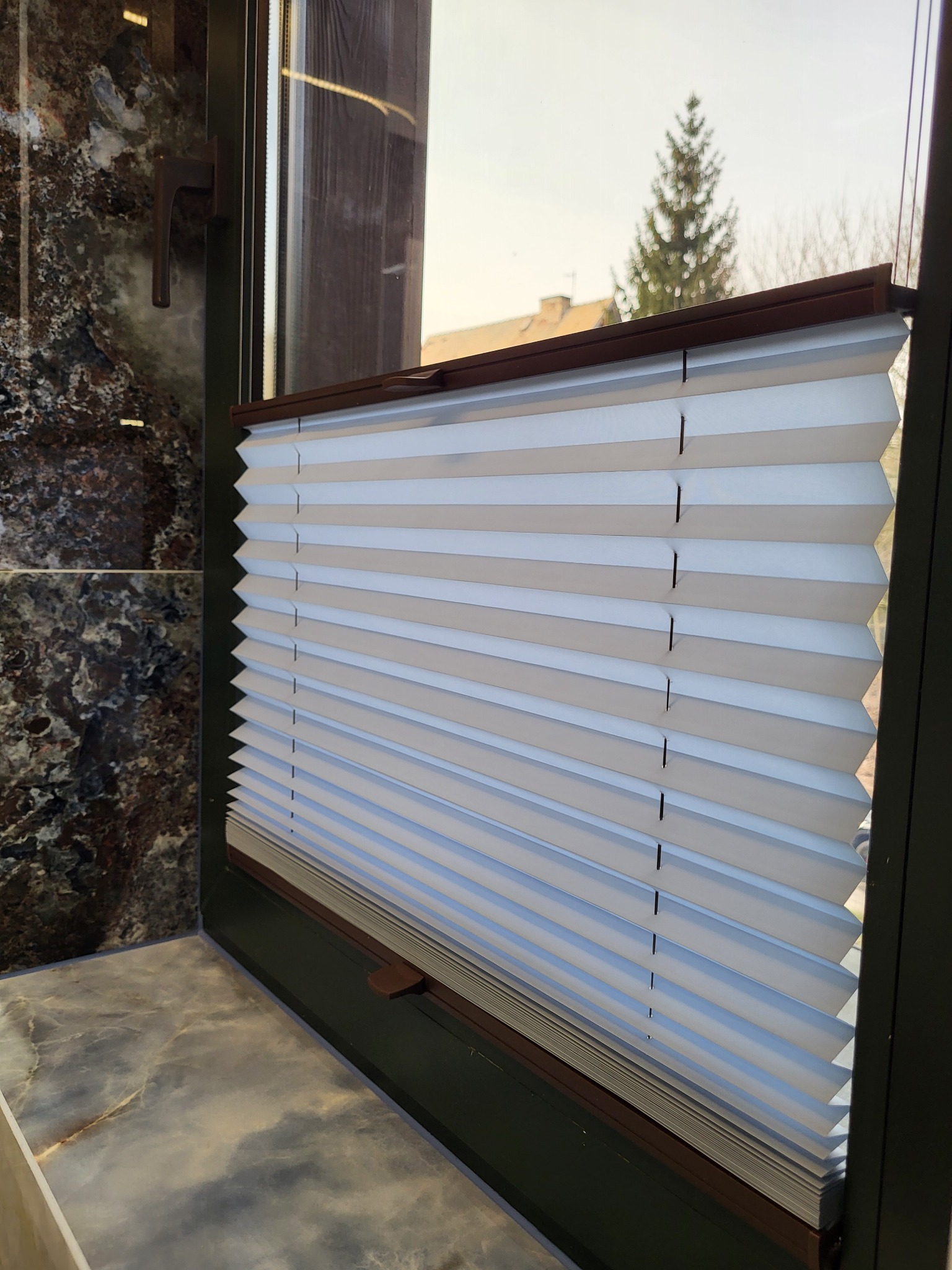 Zamknięta, biała plisa okienna z brązowymi akcentami, widoczna z boku, z widokiem na zewnątrz przez okno. Elegancki detal wykończenia okna.