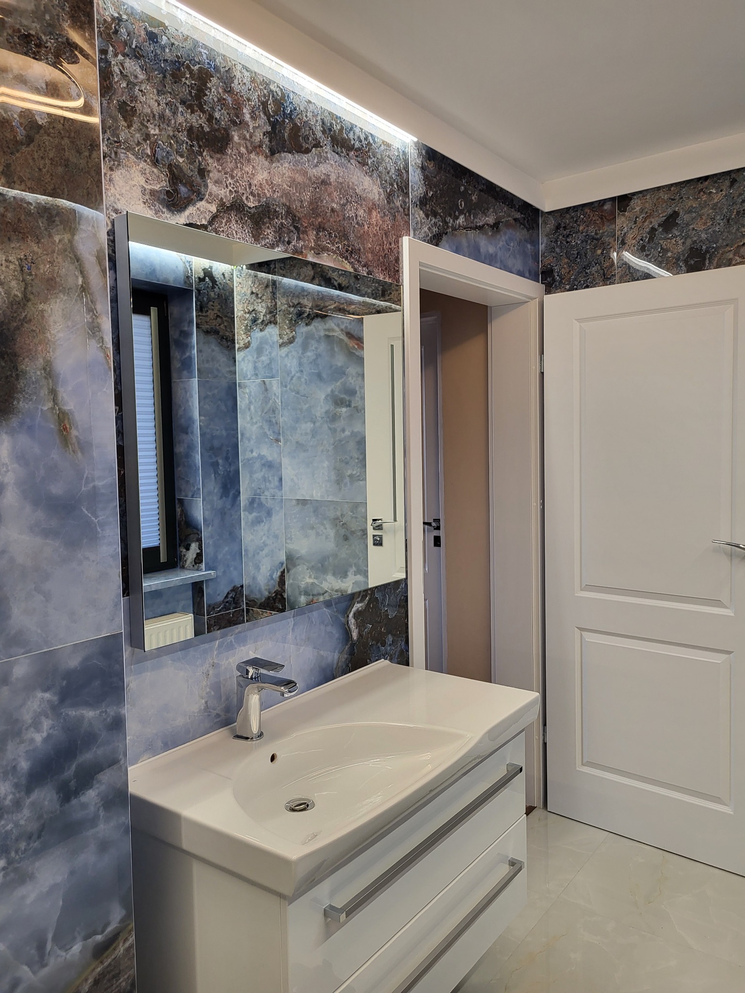 Elegancka łazienka z umywalką w szafce, lustrem z podświetleniem i roletą w oknie odbijającą się w lustrze. Płytki imitujące marmur na ścianach i podłodze.