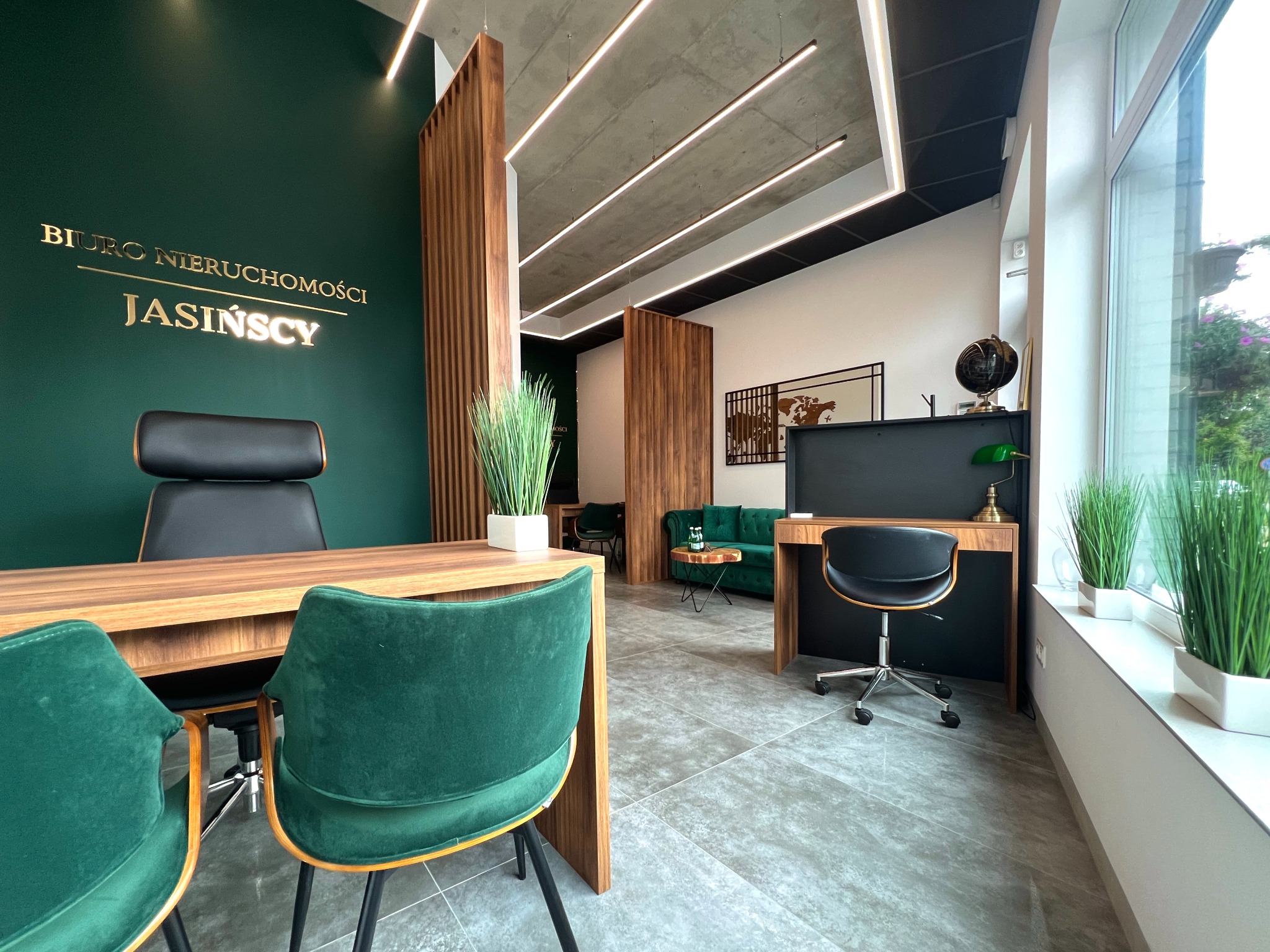 Wnętrze biura nieruchomości z zieloną ścianą, złotym napisem 'BIURO NIERUCHOMOŚCI JASIŃSCY', drewnianymi elementami, szarą podłogą, zielonymi krzesłami i biurkami