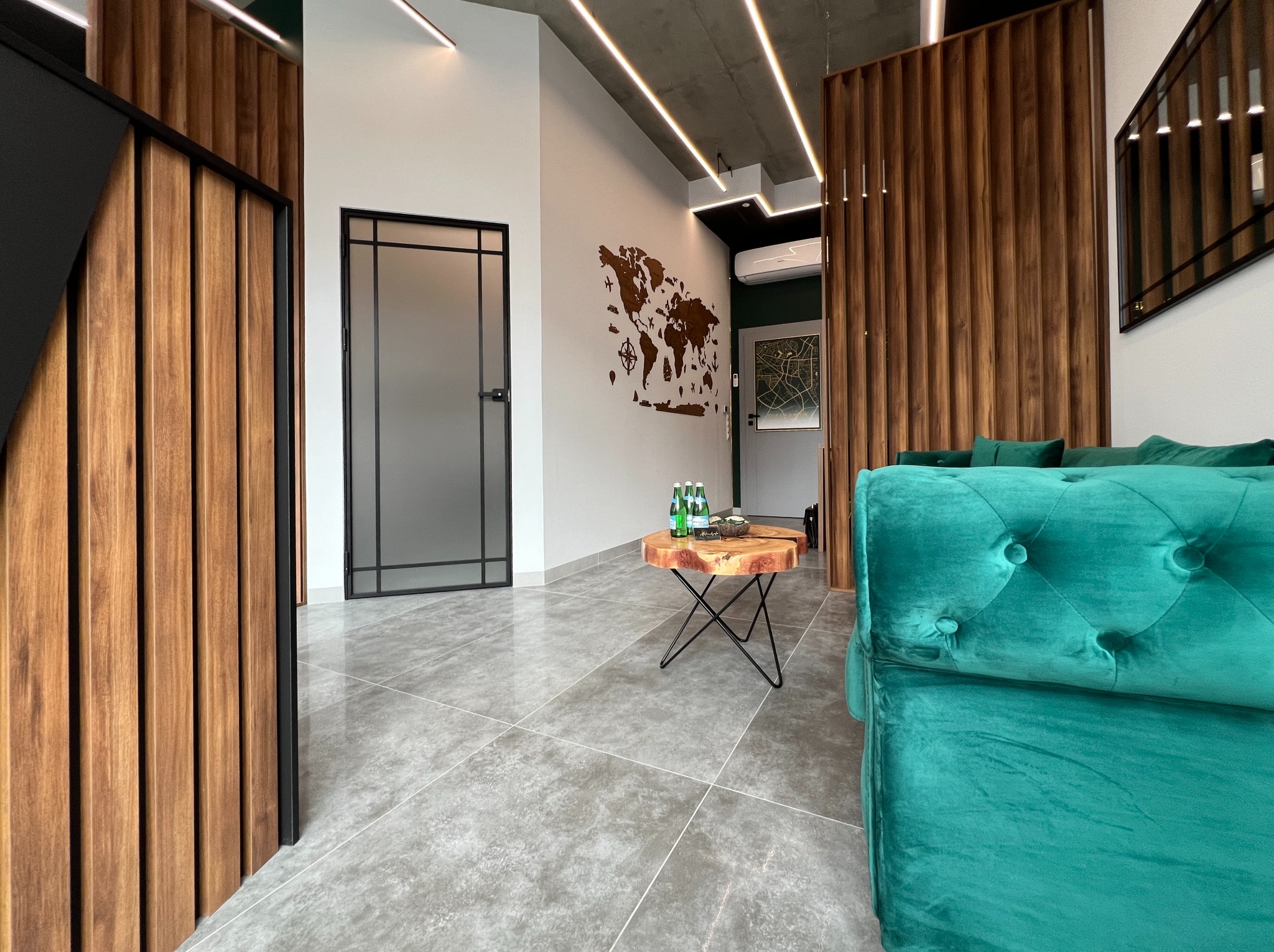 Nowoczesne wnętrze mieszkania z drewnianymi panelami, szarą podłogą i turkusową sofą; na ścianie mapa świata z drewna, stolik kawowy z plastra drewna.