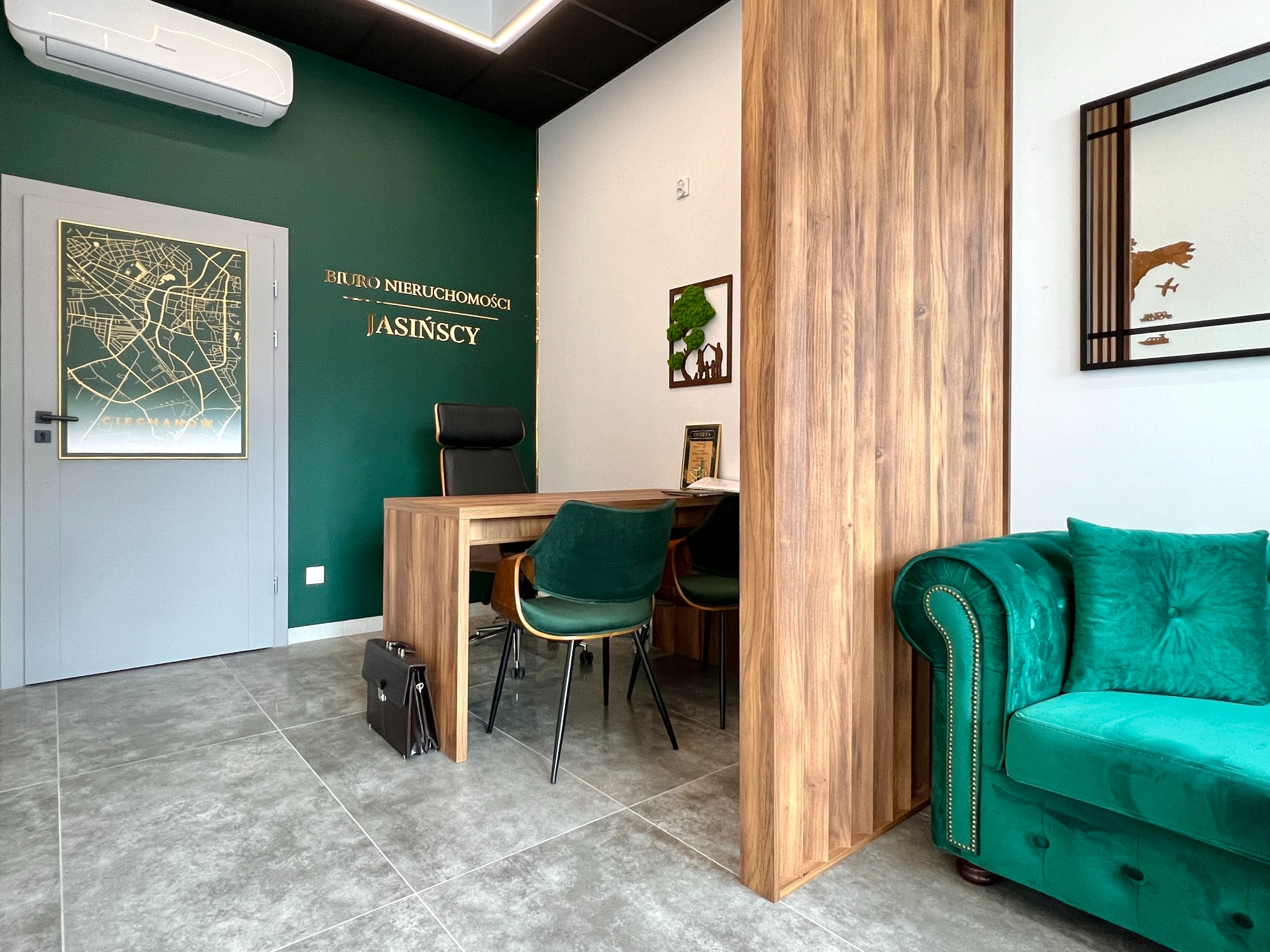 Nowoczesne biuro nieruchomości z zieloną ścianą, mapą miasta w ramie, drewnianym biurkiem, zielonymi krzesłami i sofą w stylu chesterfield, torba na dokumenty leżąca na podłodze.