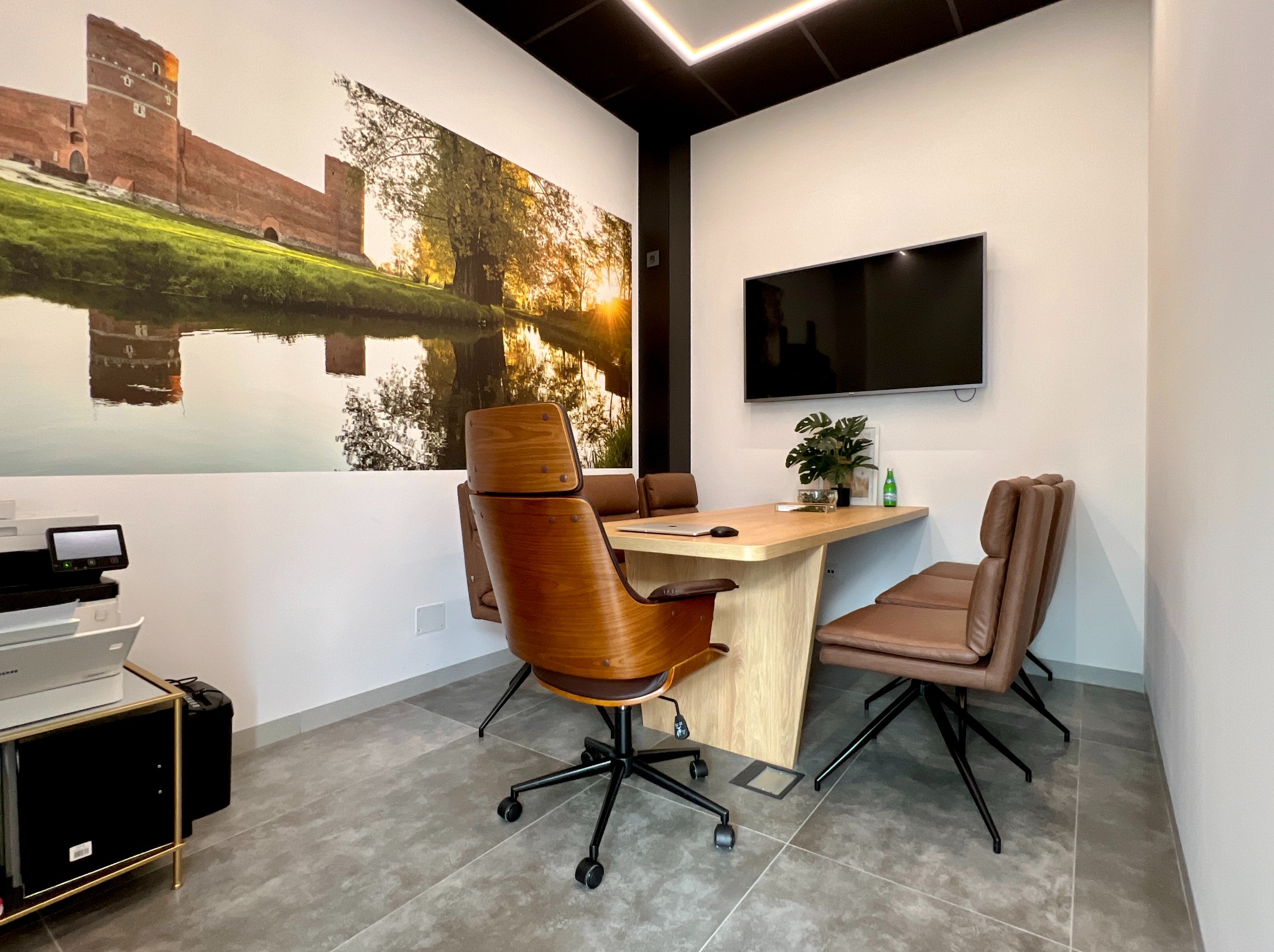 Nowoczesne biuro nieruchomości z drewnianymi krzesłami obrotowymi, jasnym biurkiem, telewizorem i fototapetą przedstawiającą zamek odbijający się w wodzie.