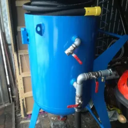 Niebieski zbiornik piaskarki z wężem, zaworami i kołami, widoczne elementy metalowe i gumowe, tło warsztatowe.