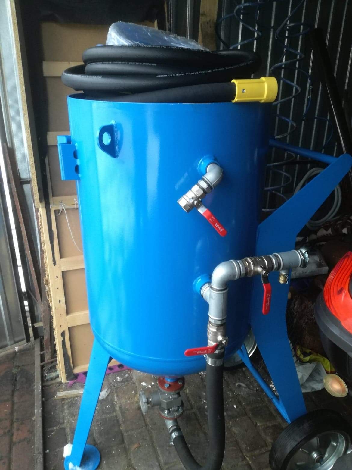 Niebieski zbiornik piaskarki z wężem, zaworami i kołami, widoczne elementy metalowe i gumowe, tło warsztatowe.