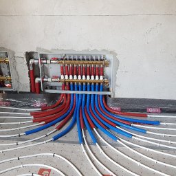 P.P.U.H. Prosper Bogusław Przybyła - Rozdzielacze ogrzewania podłogowego zamontowane w ścianie, z widocznymi rurami grzewczymi w kolorach czerwonym i niebieskim, ułożonymi na podkładzie z folią w kratkę, przed wylaniem posadzki.