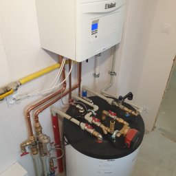 P.P.U.H. Prosper Bogusław Przybyła - Nowoczesny, biały piec gazowy marki Vaillant zamontowany na ścianie, z widocznymi miedzianymi rurami i żółtą rurą gazową, podłączony do zbiornika z licznymi zaworami i pompą obiegową, całość...
