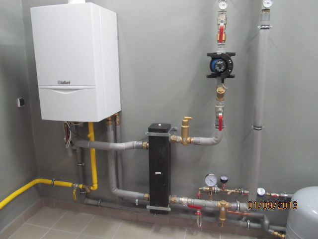 Biały piec gazowy marki Vaillant zamontowany na szarej ścianie, z widocznymi rurami w kolorze szarym i żółtym, oraz innymi elementami instalacji w pomieszczeniu z jasnymi płytkami podłogowymi.