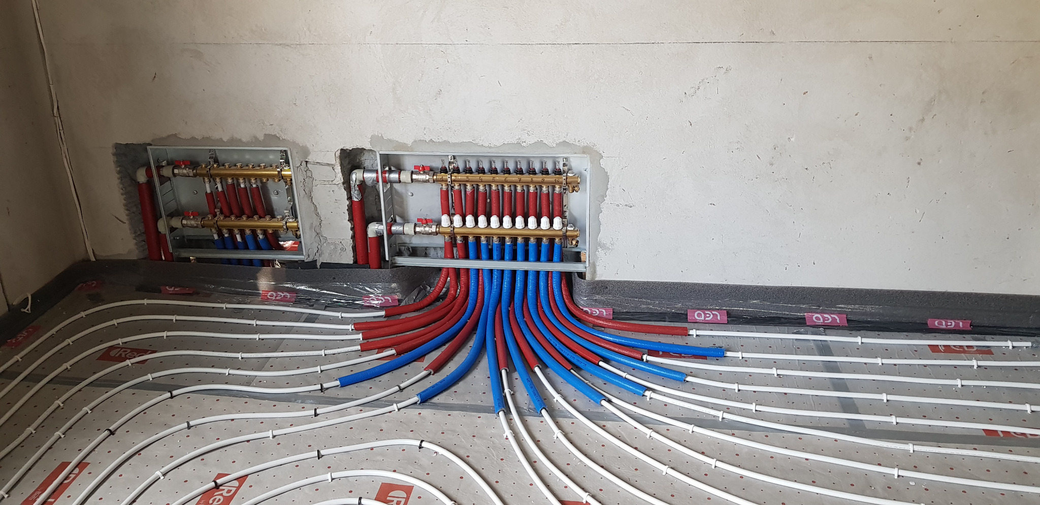 Rozdzielacze ogrzewania podłogowego zamontowane w ścianie, z widocznymi rurami grzewczymi w kolorach czerwonym i niebieskim, ułożonymi na podkładzie z folią w kratkę, przed wylaniem posadzki.