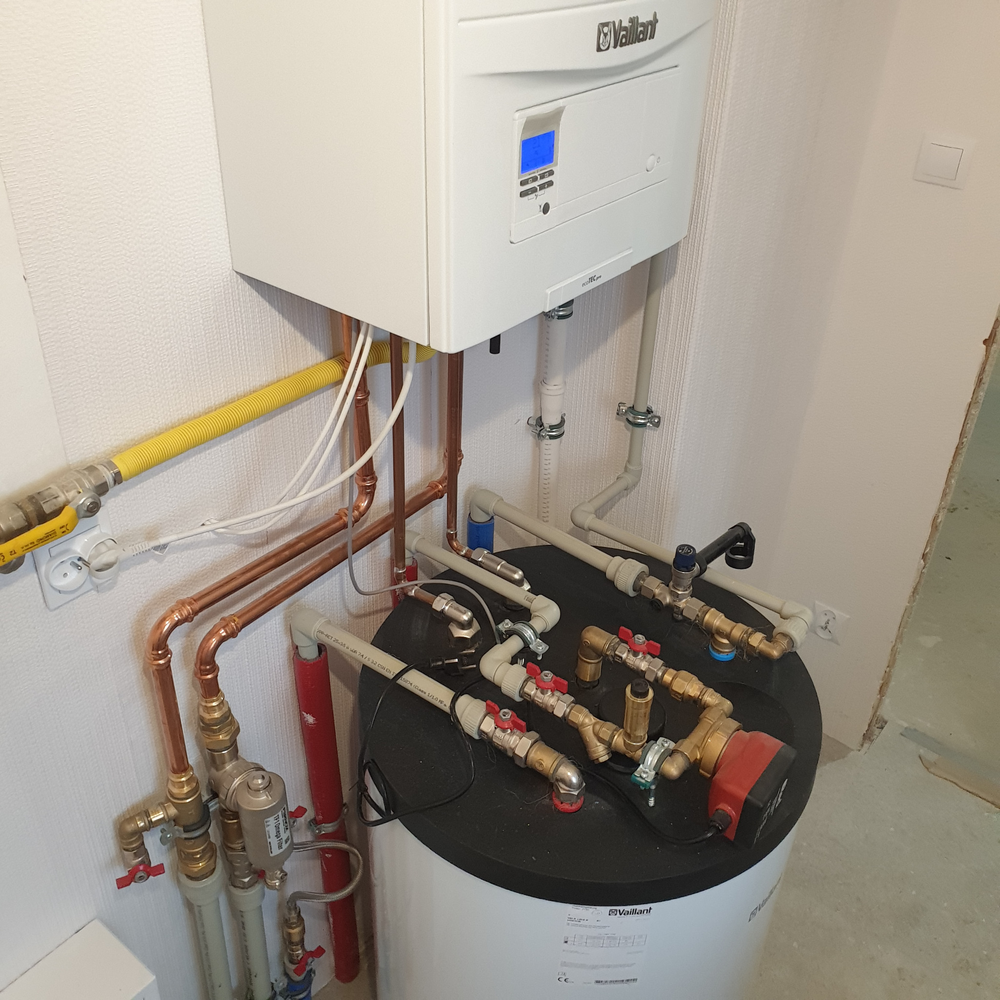 Nowoczesny, biały piec gazowy marki Vaillant zamontowany na ścianie, z widocznymi miedzianymi rurami i żółtą rurą gazową, podłączony do zbiornika z licznymi zaworami i pompą obiegową, całość...