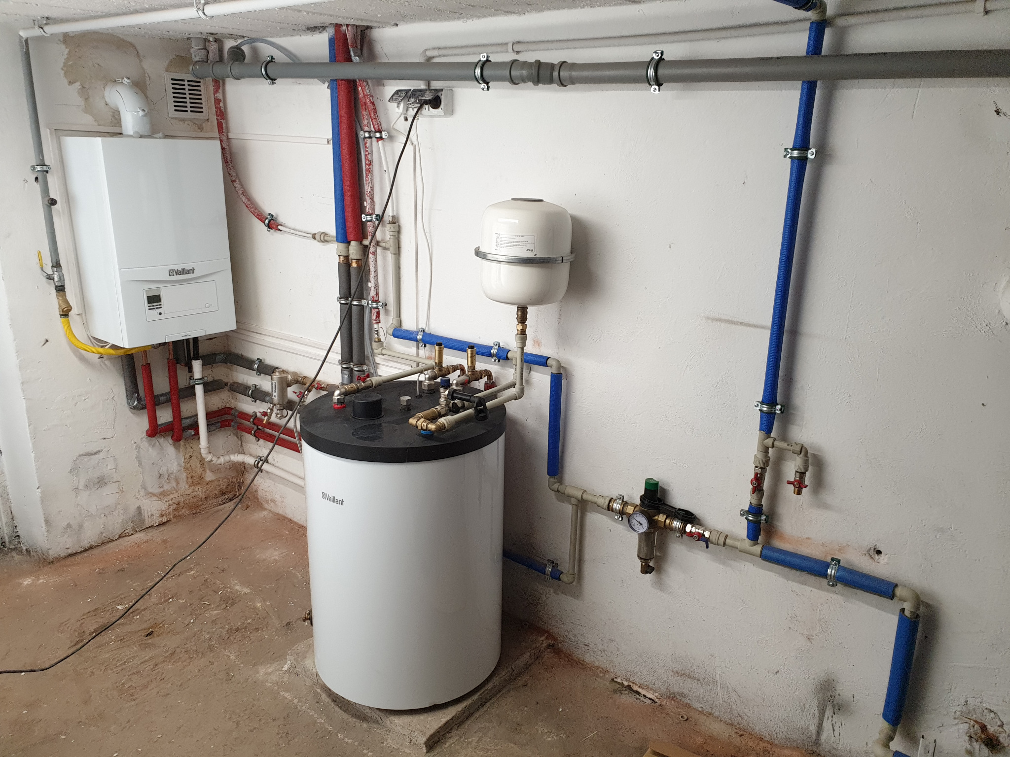 Instalacja grzewcza w piwnicy z piecem gazowym Vaillant, zbiornikiem na wodę, rurami w izolacji, manometrem i zaworami, widoczne ślady wilgoci na ścianach i podłodze.