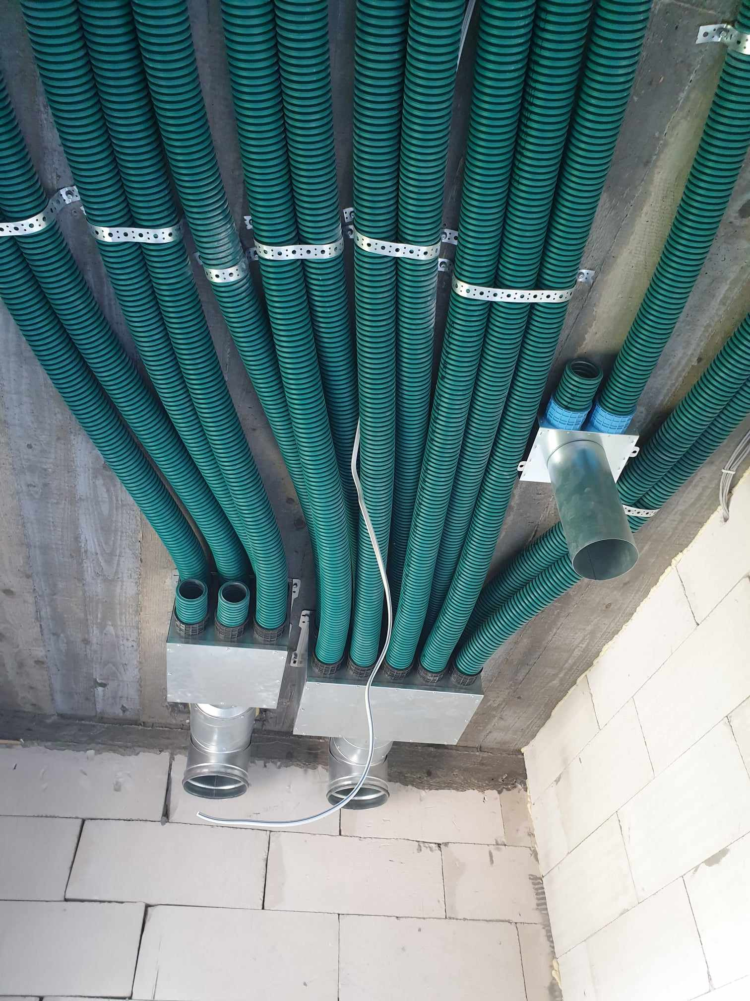 Zielone karbowane rury wentylacyjne przymocowane do sufitu za pomocą metalowych obejm, wchodzące do metalowych skrzynek i rur. Widoczne białe ściany z pustaków.