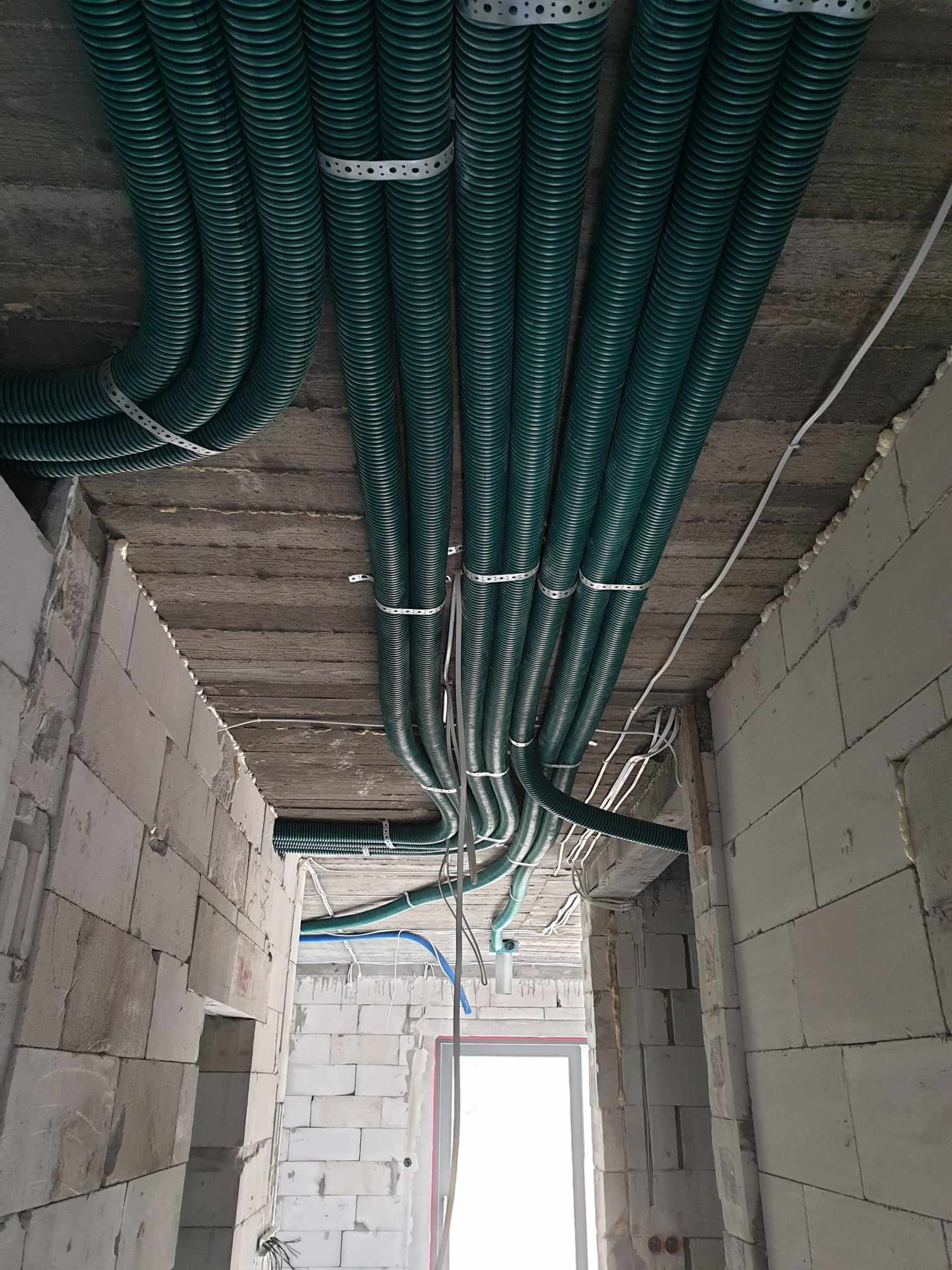 Zielone rury karbowane, przymocowane metalowymi taśmami do surowego betonowego stropu w trakcie budowy, widoczne ściany z białych bloczków.