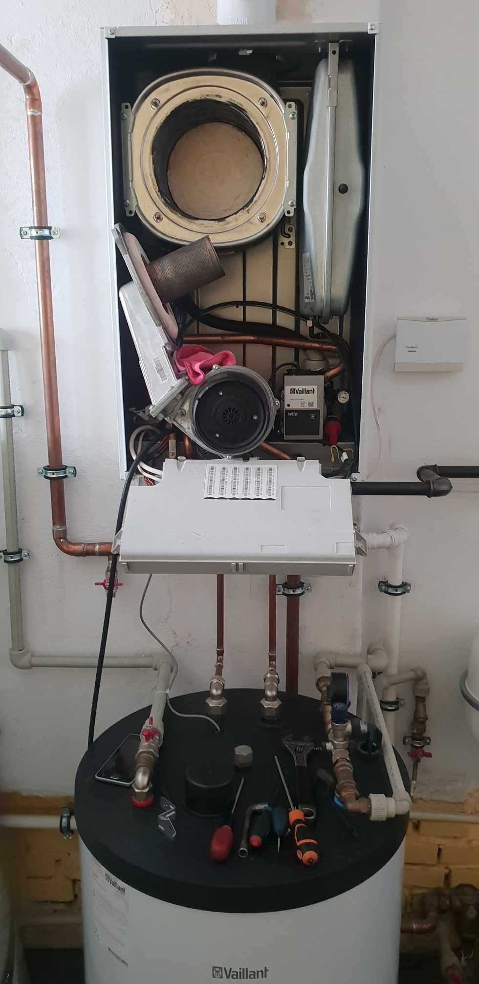 Otwarty piec gazowy Vaillant w trakcie przeglądu, widoczne elementy wewnętrzne, pod spodem zbiornik z narzędziami i telefonem komórkowym.