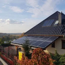 Panele słoneczne na zadaszeniu domu jednorodzinnego z czarną dachówką, widok z poziomu gruntu na tle jesiennego krajobrazu.