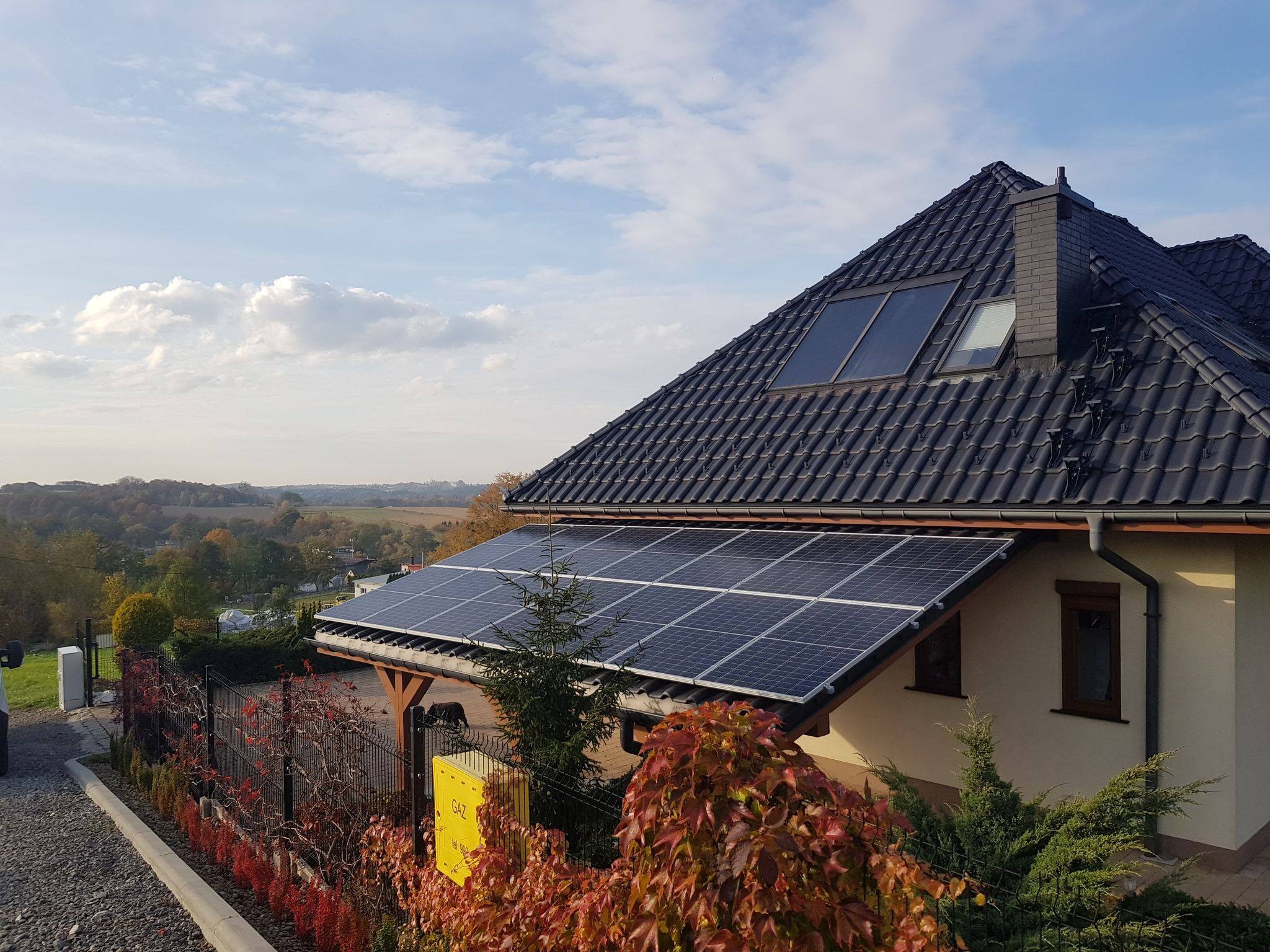 Panele słoneczne na zadaszeniu domu jednorodzinnego z czarną dachówką, widok z poziomu gruntu na tle jesiennego krajobrazu.