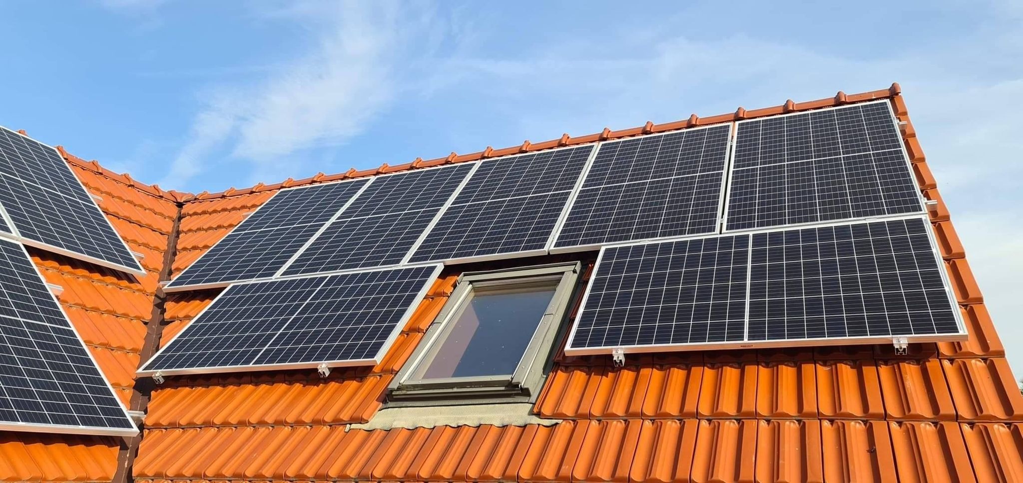 Panele słoneczne zamontowane na dachu z pomarańczowej dachówki, widoczne okno dachowe i błękitne niebo