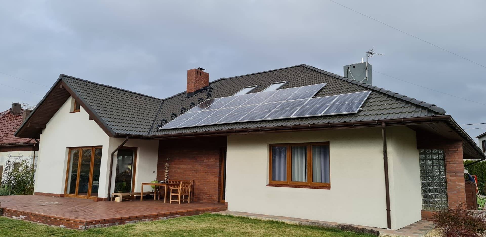 Dom jednorodzinny z zamontowanymi panelami fotowoltaicznymi na dachu z dachówki ceramicznej, widoczne okna dachowe, komin z cegły, taras z cegły klinkierowej i meble ogrodowe.