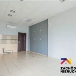 Puste pomieszczenie z beżowymi płytkami podłogowymi i częściowo wykończoną ścianą z rurami, klimatyzator na ścianie, widoczne ślady po wcześniejszych pracach.