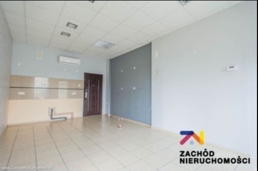 Puste pomieszczenie z beżowymi płytkami podłogowymi i częściowo wykończoną ścianą z rurami, klimatyzator na ścianie, widoczne ślady po wcześniejszych pracach.