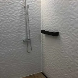 Nowoczesna kabina prysznicowa z geometrycznymi białymi płytkami 3D na ścianach i drewnopodobną podłogą, wyposażona w deszczownicę i czarną półkę.