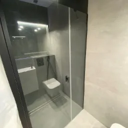 Nowoczesna łazienka w odcieniach szarości z kabiną prysznicową typu walk-in, minimalistyczną umywalką i podwieszanym WC, zrealizowana w mieszkaniu w Miechowie.