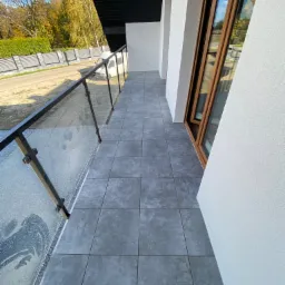 Szare płytki na balkonie z przeszkloną balustradą i drewnianą ramą okienną, widok na ulicę z drzewami w tle.