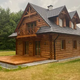 Drewniany dom z bali z czarnym dachem i tarasem, otoczony zielenią, w tle las i pole.