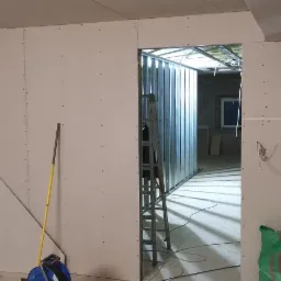 Montaż płyt gipsowo-kartonowych na poddaszu, widoczne profile metalowe, drabina, narzędzia oraz worki budowlane.