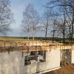 Wznoszenie ścian parteru domu murowanego z betonu komórkowego, z widocznym deskowaniem wieńca, na tle jesiennego krajobrazu z drzewami bez liści.