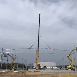 Montaż stalowej konstrukcji dachu hali przemysłowej przy użyciu dźwigu i podnośników koszowych; widoczni pracownicy w kamizelkach odblaskowych.