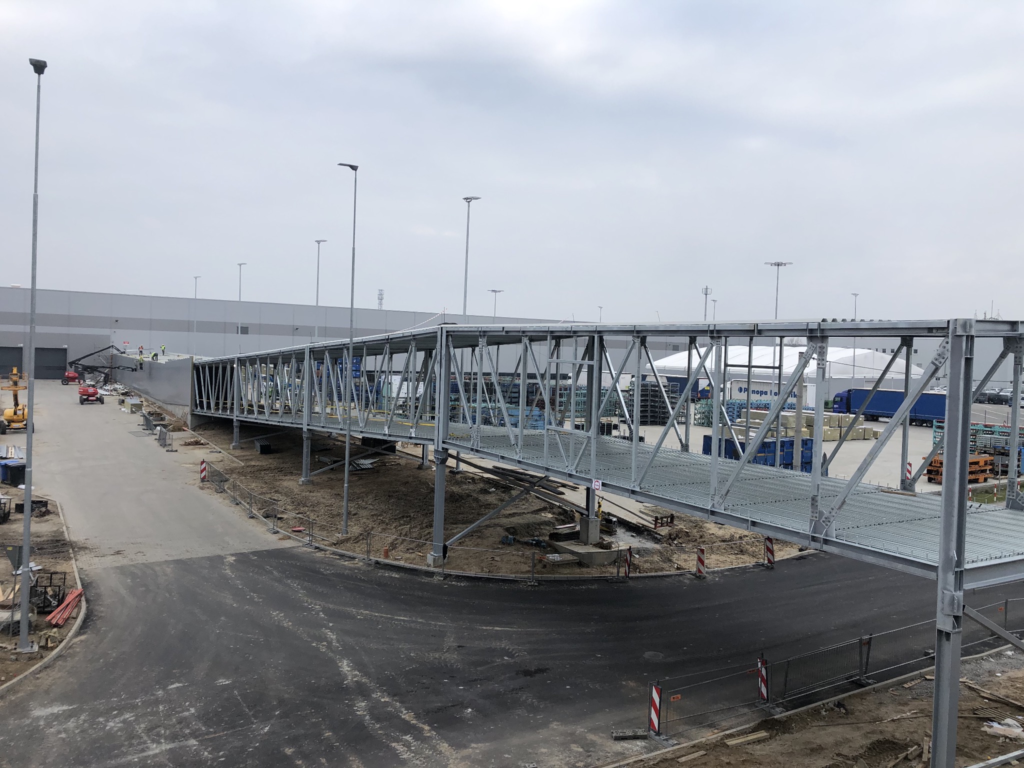 Nowo wybudowana, ocynkowana kładka dla pieszych nad drogą do hali magazynowej, widok z góry na plac budowy i robotników w oddali.