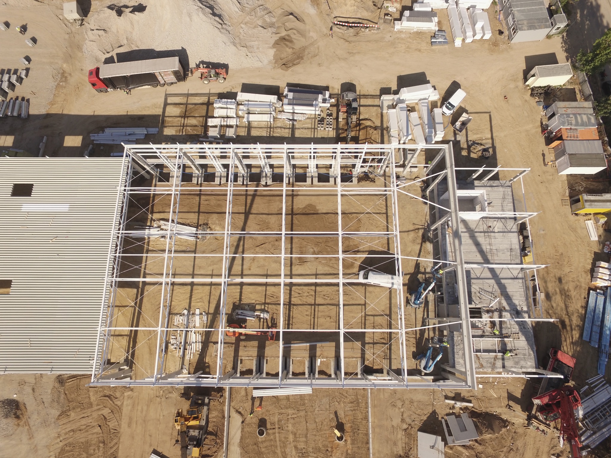 Zdjęcie z lotu ptaka przedstawia budowę hali z białą stalową konstrukcją szkieletową, częściowo pokrytą blachą falistą; widoczne maszyny budowlane, materiały składowane na placu oraz fundamenty...