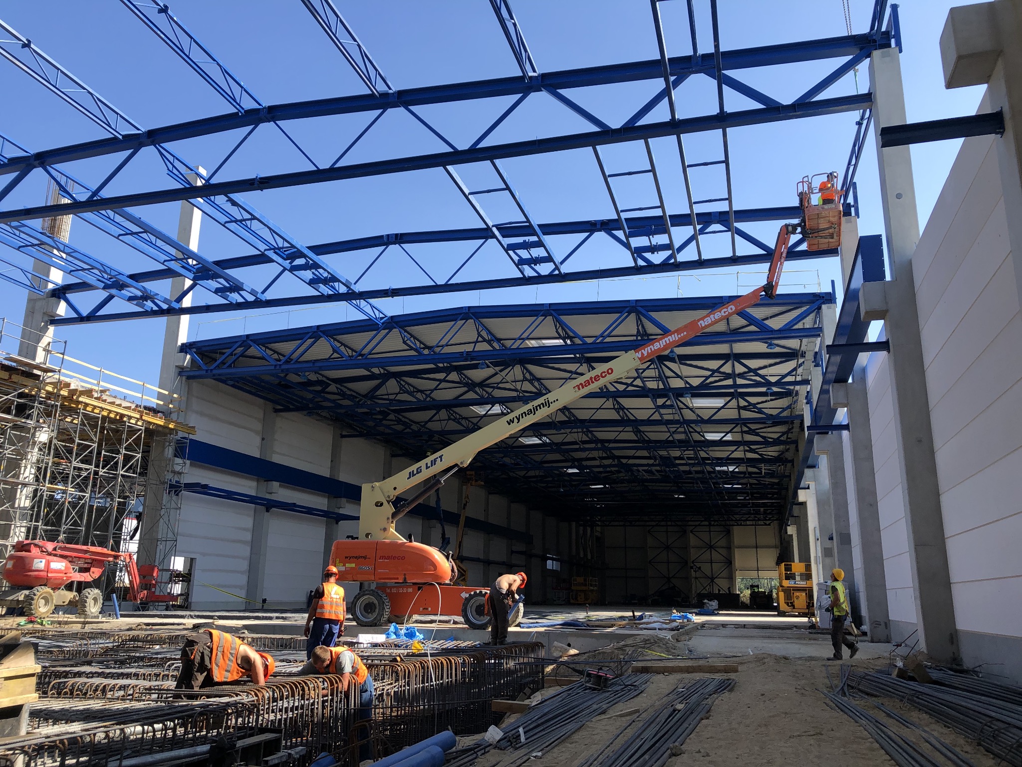 Montaż niebieskiej konstrukcji stalowej hali przez pracowników w pomarańczowych kamizelkach, widoczny podnośnik JLG i zbrojenie fundamentów.