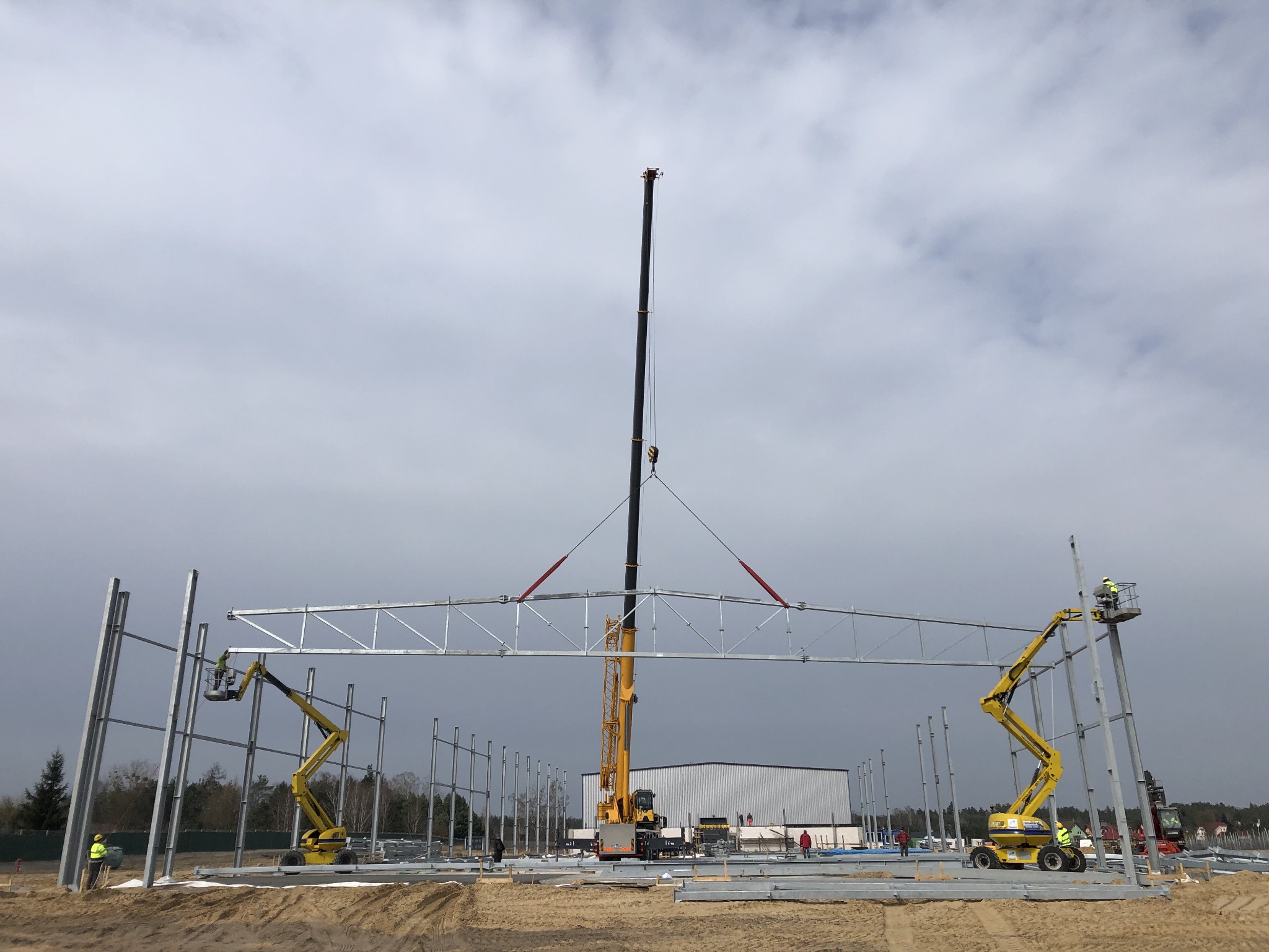 Montaż stalowej konstrukcji dachu hali przemysłowej przy użyciu dźwigu i podnośników koszowych; widoczni pracownicy w kamizelkach odblaskowych.