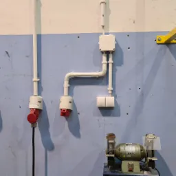 Ściana warsztatu z widocznymi elementami instalacji elektrycznej: gniazda przemysłowe z czerwonymi wtykami, wyłącznik obrotowy, puszka instalacyjna z peszlem, podwójny włącznik; na dole szlifierka...