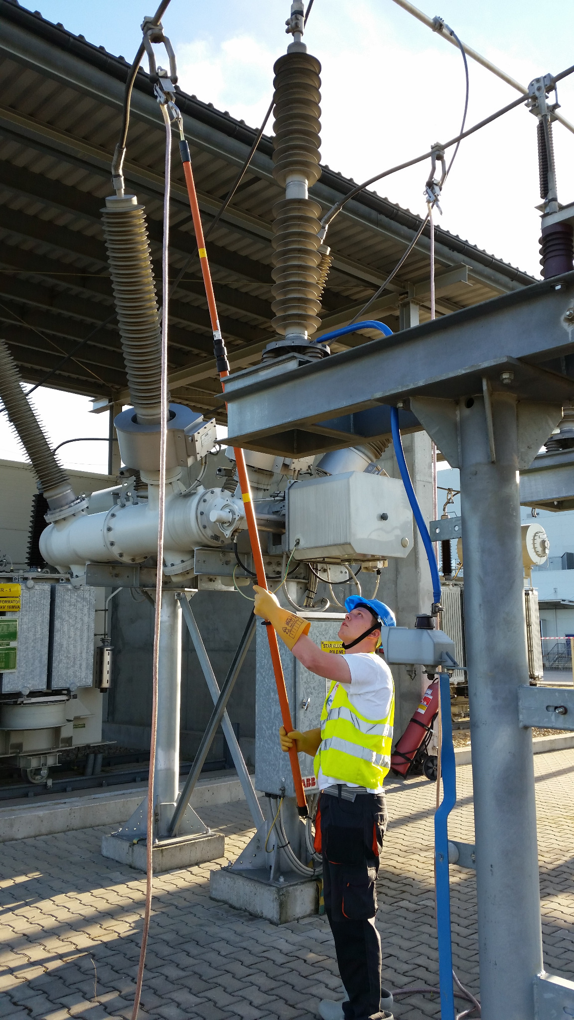 Elektryk w kasku i kamizelce odblaskowej, używający tyczki izolacyjnej przy transformatorze wysokiego napięcia na zewnątrz budynku w słoneczny dzień.